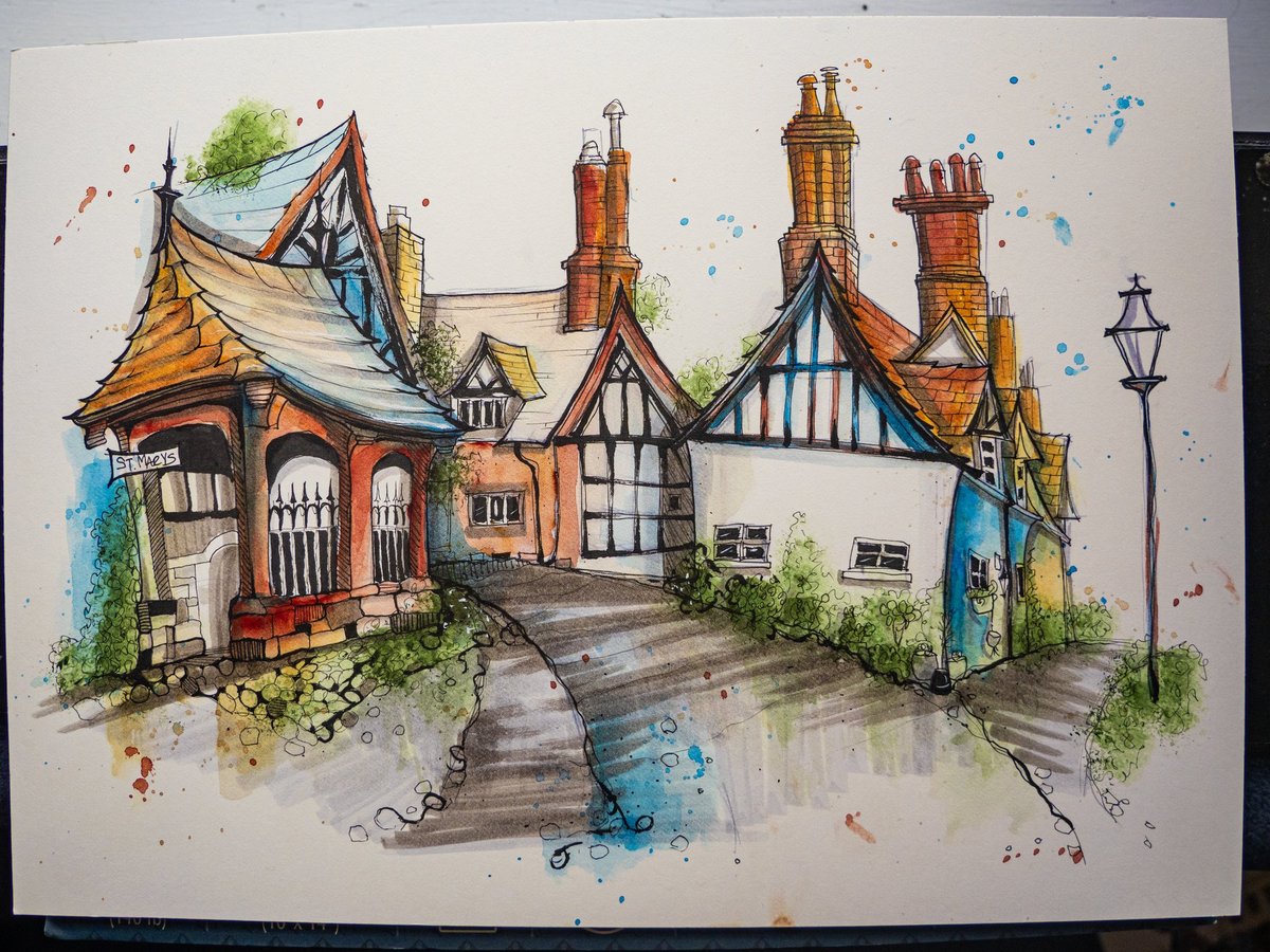 A detailed urban sketch of a whimsical Tudor-style row of buildings, featuring distinctive orange-red brick structures with dark timber framing, steep pitched roofs with chimneys, and arched Gothic windows. The composition shows a village street scene rendered in ink pen with watercolor washes in blues, greens, and warm earth tones. A decorative street lamp stands to the right, and creative splatter details in blue and rust-colored watercolor add artistic flair to the foreground and background. The left building features a sign reading "St. Marys." The sketch captures charming English architectural character with lush green foliage sprouting from the buildings.