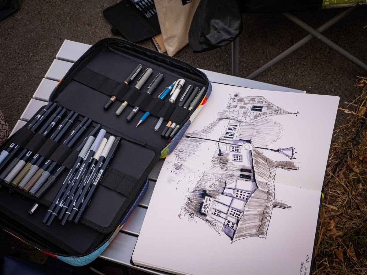 A overhead flat-lay composition of urban sketching supplies and student work displayed outdoors. A professional black marker/pen case is open on the left, revealing dozens of organized fine-tip pens, markers, and colored pencils in grayscale and accent colors. To the right, a white sketchbook displays detailed architectural line drawings of historic street buildings with decorative cross-hatching and linework in black ink. The scene captures an active sketching session or workshop setup on textured ground, with folding chairs and personal items visible in the background.
