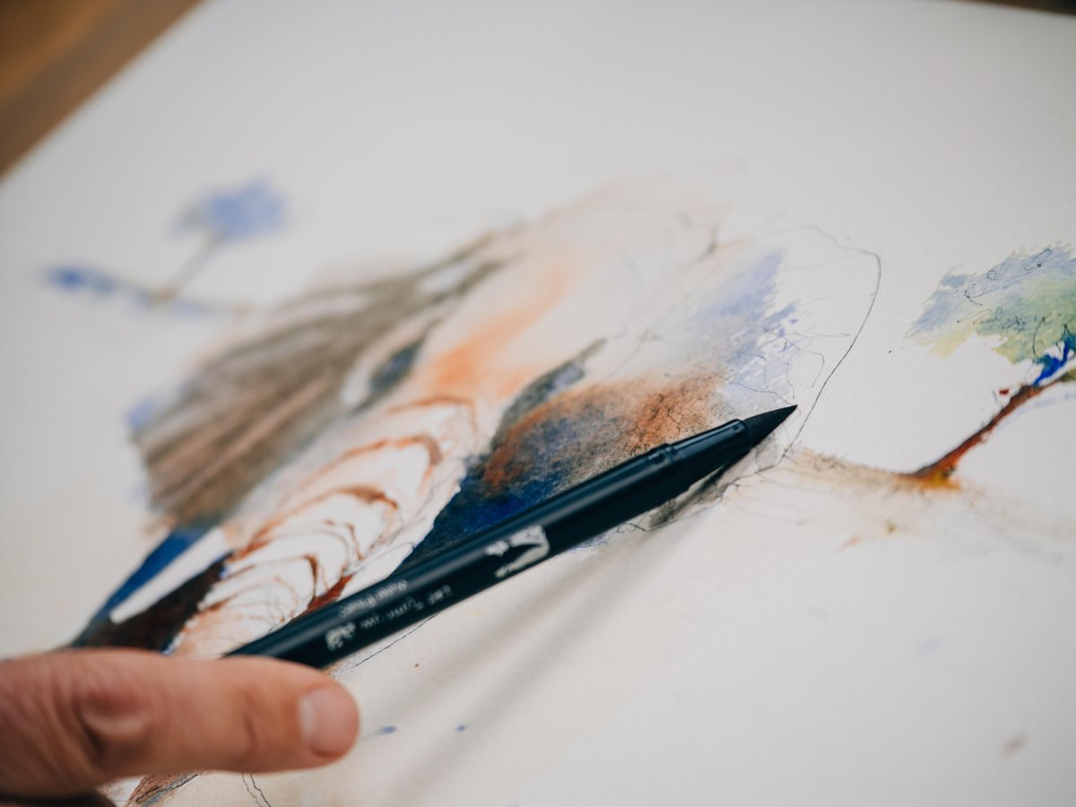 A close-up photograph of a hand holding a watercolor brush mid-stroke over a detailed watercolor sketch. The artwork features landscapes and wildlife rendered in soft washes of blue, orange, brown, and green tones. The brush is dark-handled with blue bristles, actively applying color to the paper. The composition captures the intimate moment of artistic creation, with blurred background sketches visible and layers of wet paint visible on the paper surface.