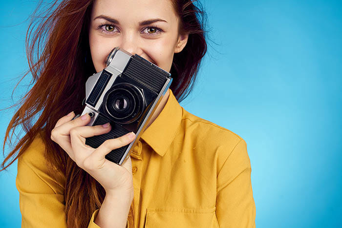 portrait of a lady in a yellow blouse against a blue background holding a camera and smiling
