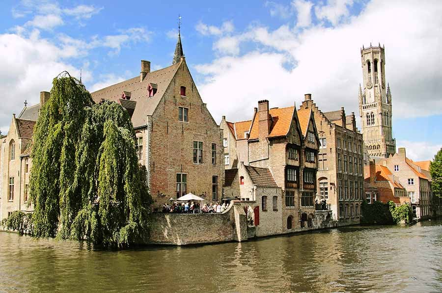 A canal in Bruges lined with brick gabled houses, with the Belfry tower rising behind them