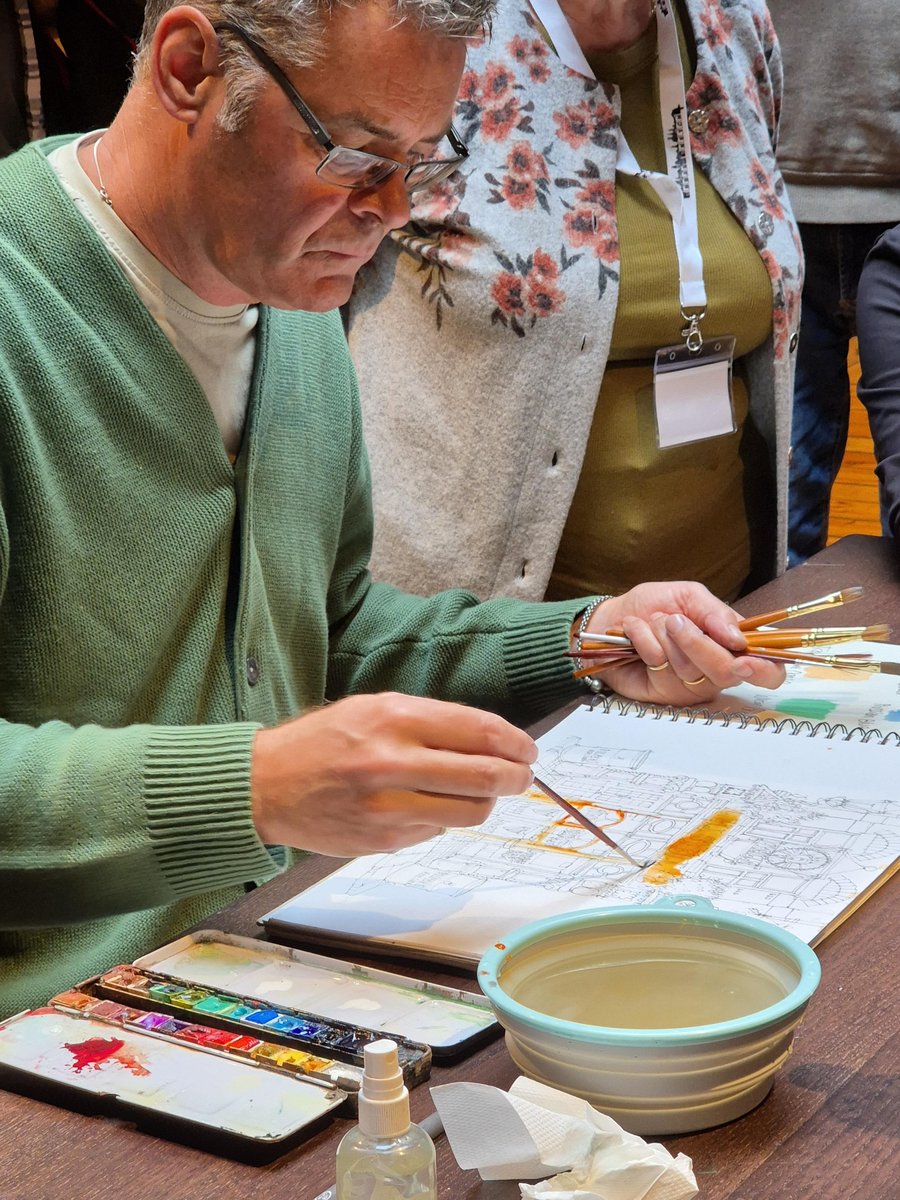 A middle-aged man in a green knit sweater wearing rectangular-framed glasses concentrates intently on a detailed architectural sketch in his spiral-bound notebook, holding multiple paintbrushes. A water bowl, paint palette with bright pigments, and fellow course participants are visible at a wooden table during what appears to be an indoor sketching workshop or class session. The warm, focused lighting and collaborative atmosphere suggest an active learning environment.