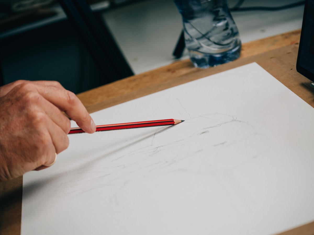 A close-up shot of hands holding a red pencil over a white sketch pad on a wooden table. Light sketch marks are visible on the paper. In the blurred background, another person wearing blue jeans is partially visible, suggesting an outdoor or workshop setting with natural daylight. The scene captures an active sketching demonstration or instruction moment.