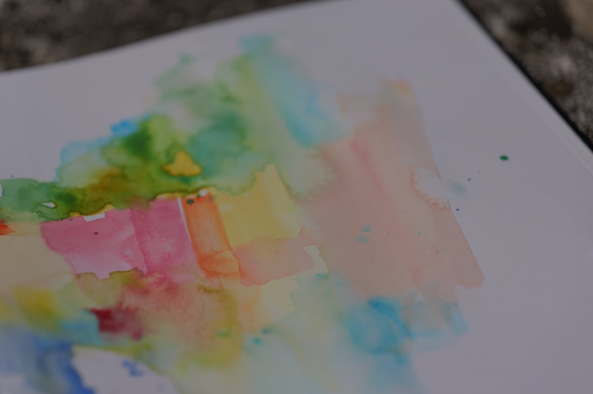 A close-up photograph of a watercolor sketch in progress on white paper, featuring soft washes of pink, yellow, green, blue, and peachy tones blending together. The composition shows abstract gestural marks and wet color bleeding effects, characteristic of watercolor painting technique. The artwork is photographed from above at an angle, with outdoor ground/stone visible in the background, suggesting this was captured during an outdoor sketching session.