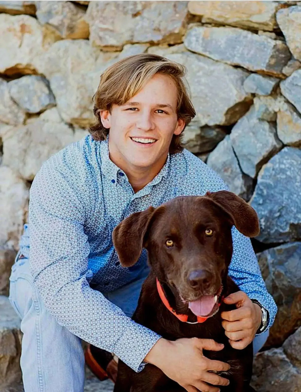 Timberline High senior in light blue shirt sitting with chocolate Labrador in front of a stone wall, both smiling at the camera