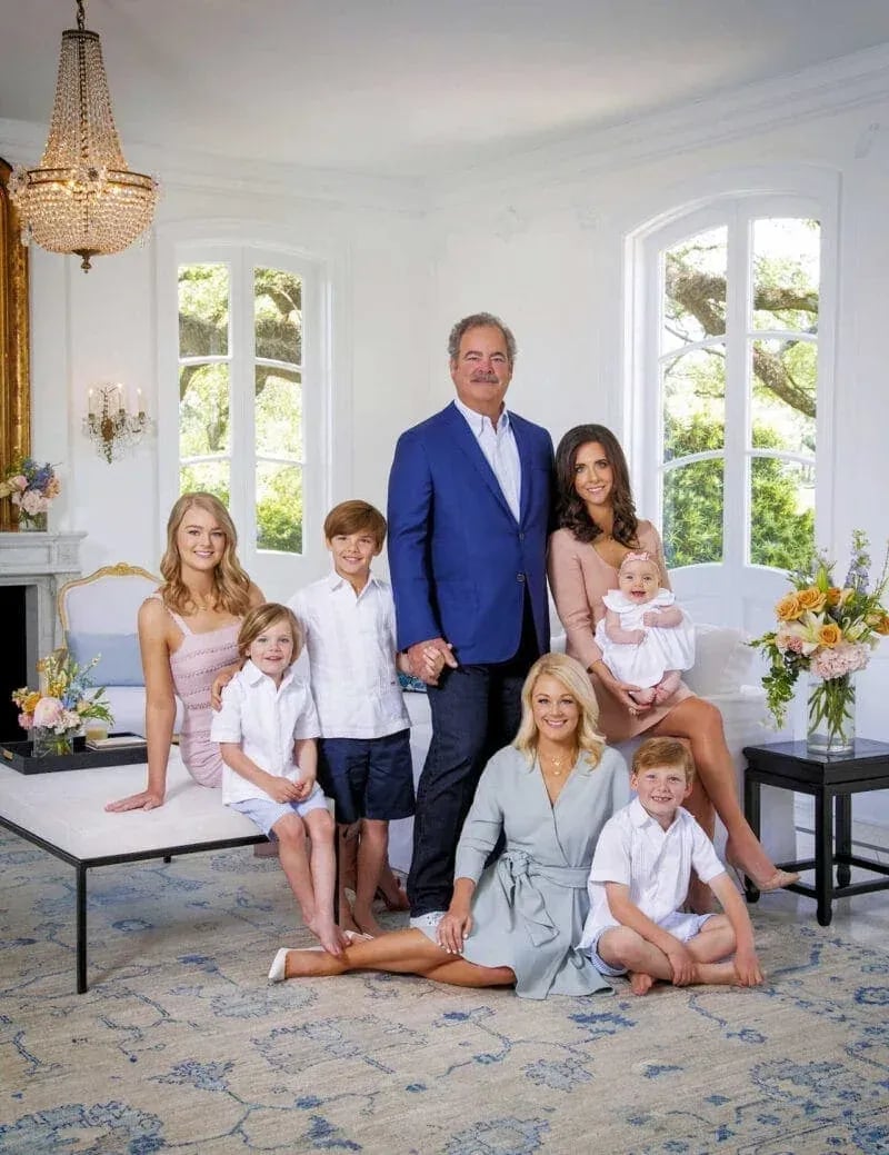 Indoor family portrait with eight individuals in semi-formal attire, including three adult women, one man, three children, and a baby, seated in an elegant room with chandelier, fireplace, and floral decor—part of Idaho Photography Studios’ Masters Family Portraits series.