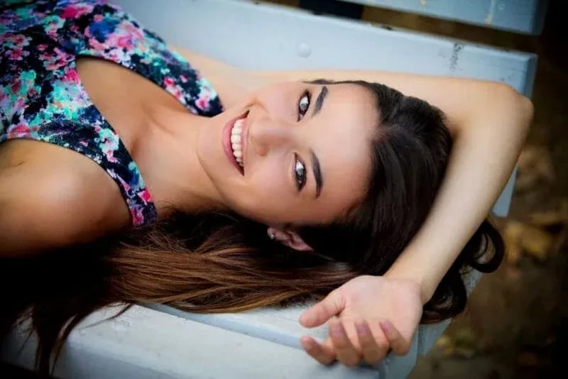Idaho high school senior with long brown hair smiling on a wooden bench outdoors, wearing a colorful floral sleeveless top in a natural setting.