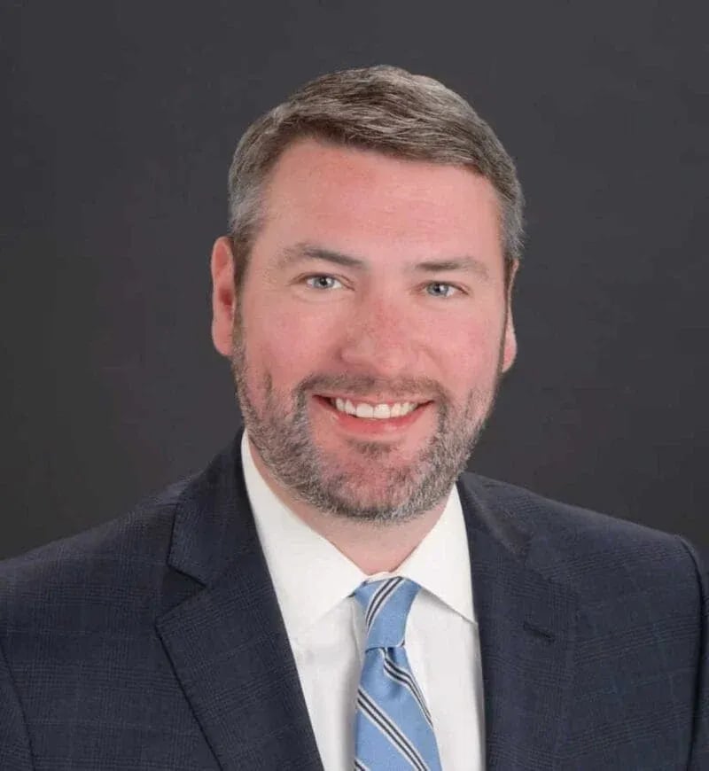 Professional headshot of a smiling man in a dark suit, white shirt, and light blue striped tie, posed against a plain dark background—photographed by Idaho Photography Studios.