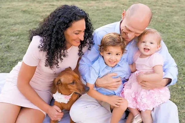 Outdoor family lifestyle portrait featuring two adults, two children, and a brown dog with a flower collar, seated on a blanket in a grassy area—photographed by Idaho Photography Studios.