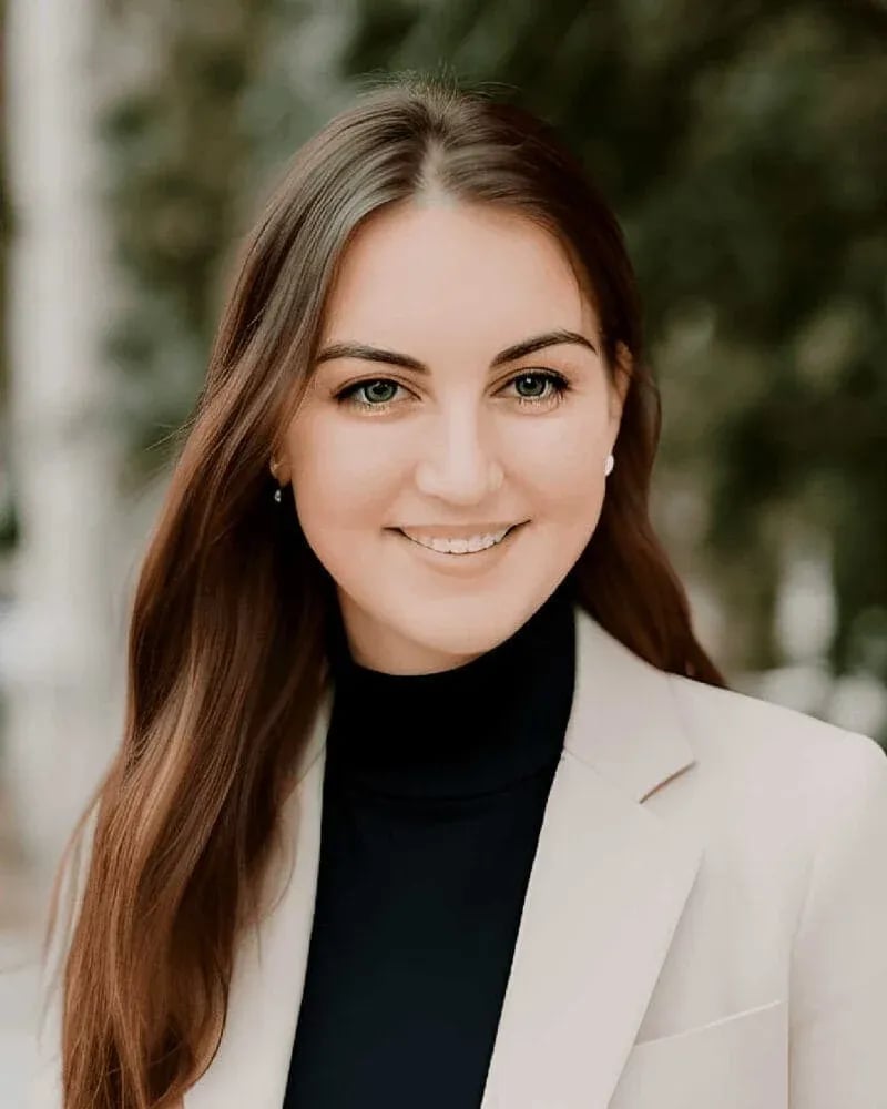 Centennial High senior in light blazer and black turtleneck standing outdoors with greenery in the background, gazing confidently