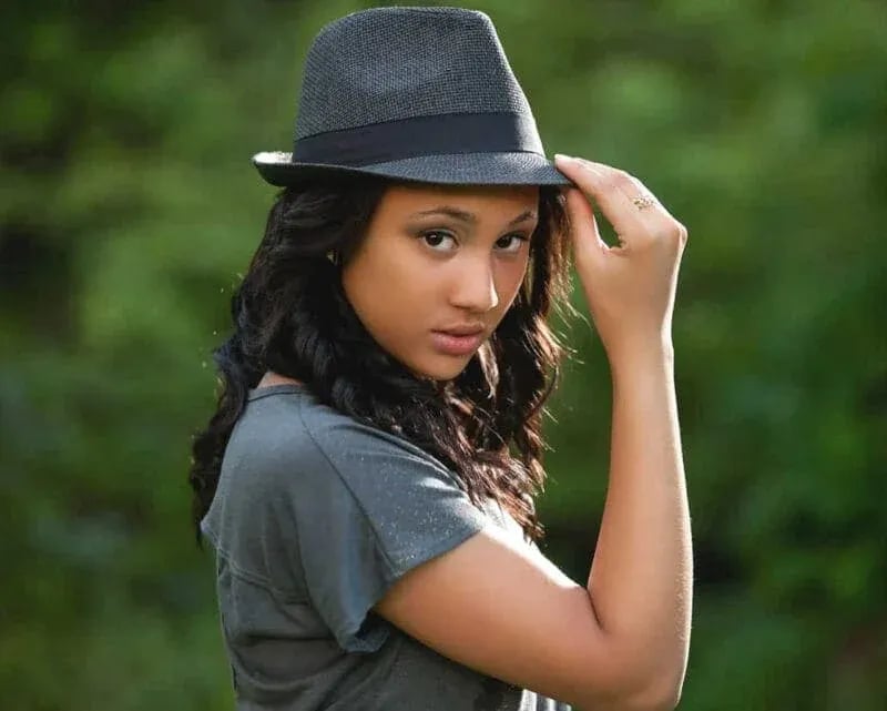 Columbia High senior in gray t-shirt and fedora smiling outdoors with green foliage background
