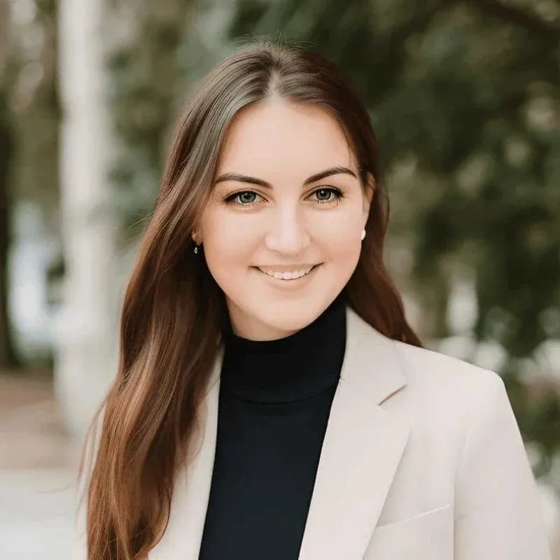 Professional outdoor portrait of a woman with long brown hair, wearing a beige blazer over a black turtleneck, standing in front of greenery.