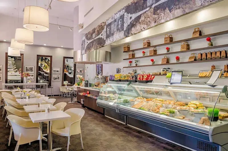 Interior of a modern café in Caldwell, Idaho, featuring pendant lighting, white cabinetry, dining tables, and a refrigerated display case filled with food items.