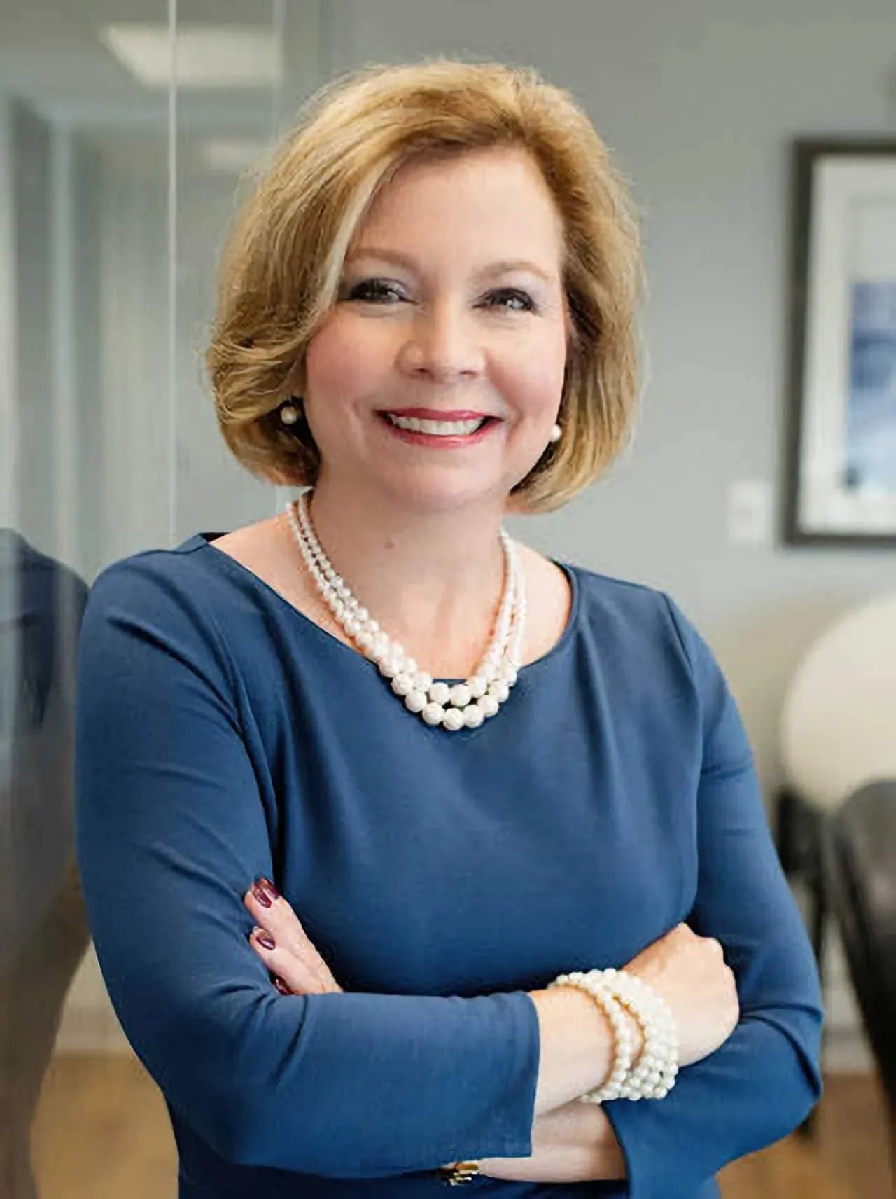 Professional headshot of a smiling woman with short blonde hair, wearing a blue dress and pearl accessories in an office setting.