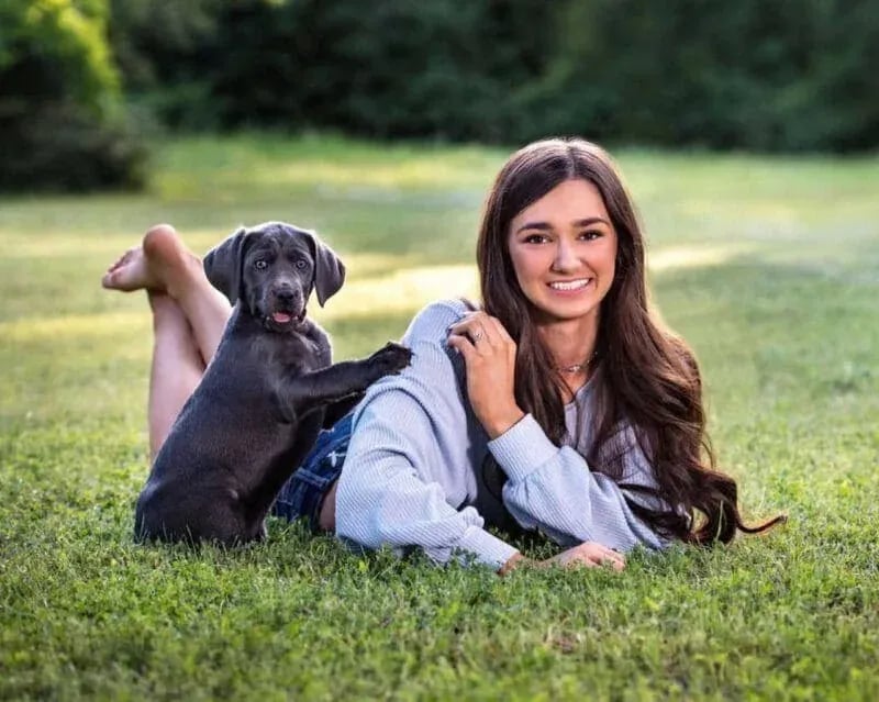 Mountain Home High senior lying on grass with a small puppy, smiling in a sunlit park setting