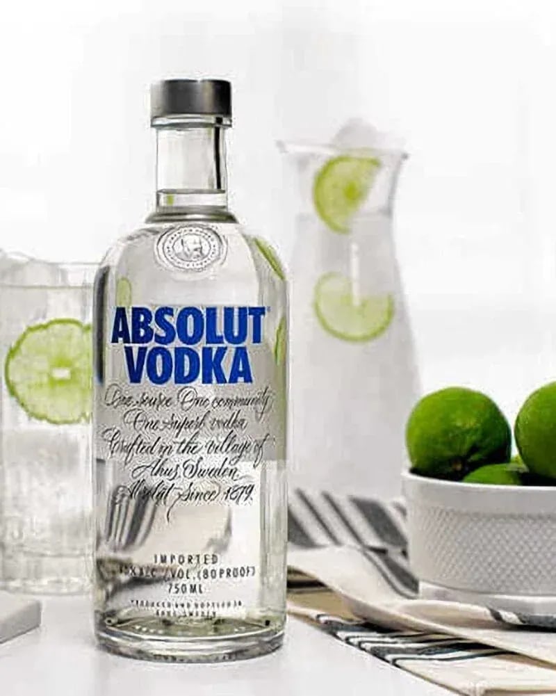 Absolut Vodka bottle surrounded by three tall glasses filled with ice, lime slices, and clear liquid, styled with a striped cloth, hexagonal coasters, and a bowl of limes on a modern tabletop.