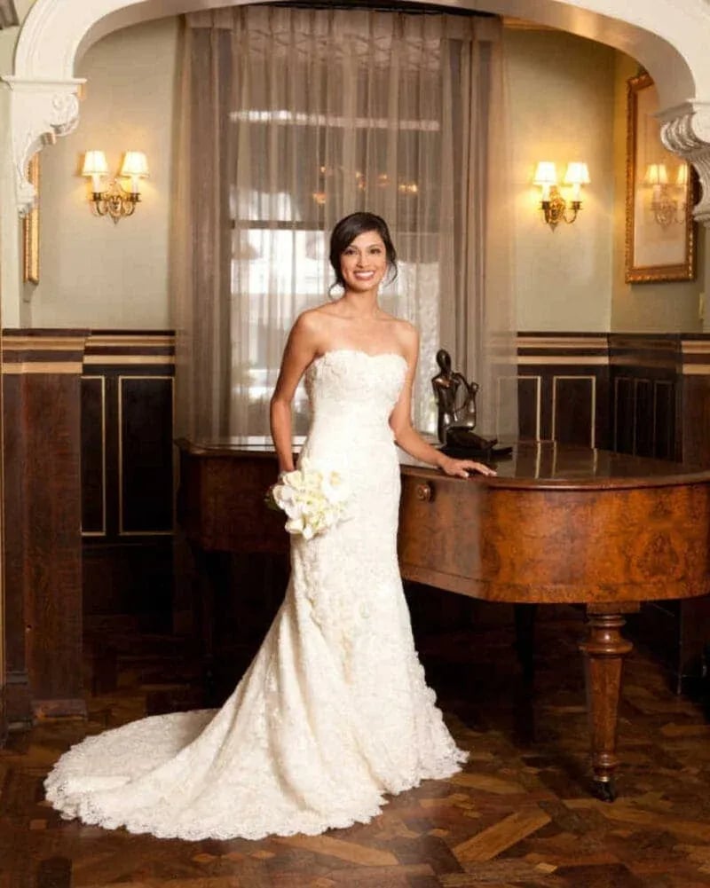 Bride in strapless white lace wedding gown with long train, holding bouquet beside grand piano in elegant room.