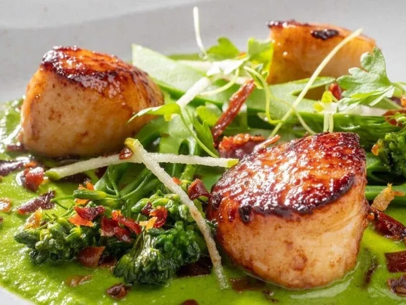 Gourmet plated scallops with golden crust on green puree, garnished with broccolini, apple slices, bacon bits, and arugula.