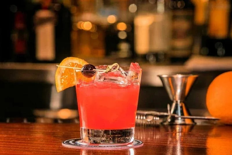 Vibrant red cocktail served in a short glass with ice, garnished with orange slice and cherries, on a polished wooden bar.