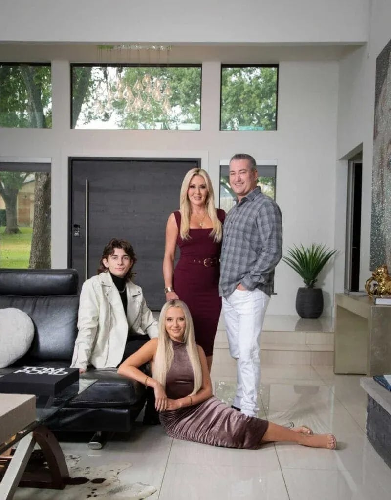 : Stylish family portrait of four people in a modern living room with large windows, black leather sofa, and contemporary decor.