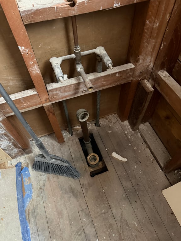 Exposed bathroom wall: original galvanized supply lines and a three-valve tub assembly mounted between studs, a rectangular cut in the dusty floorboards for the drain, a grey broom leaning on the left stud.
