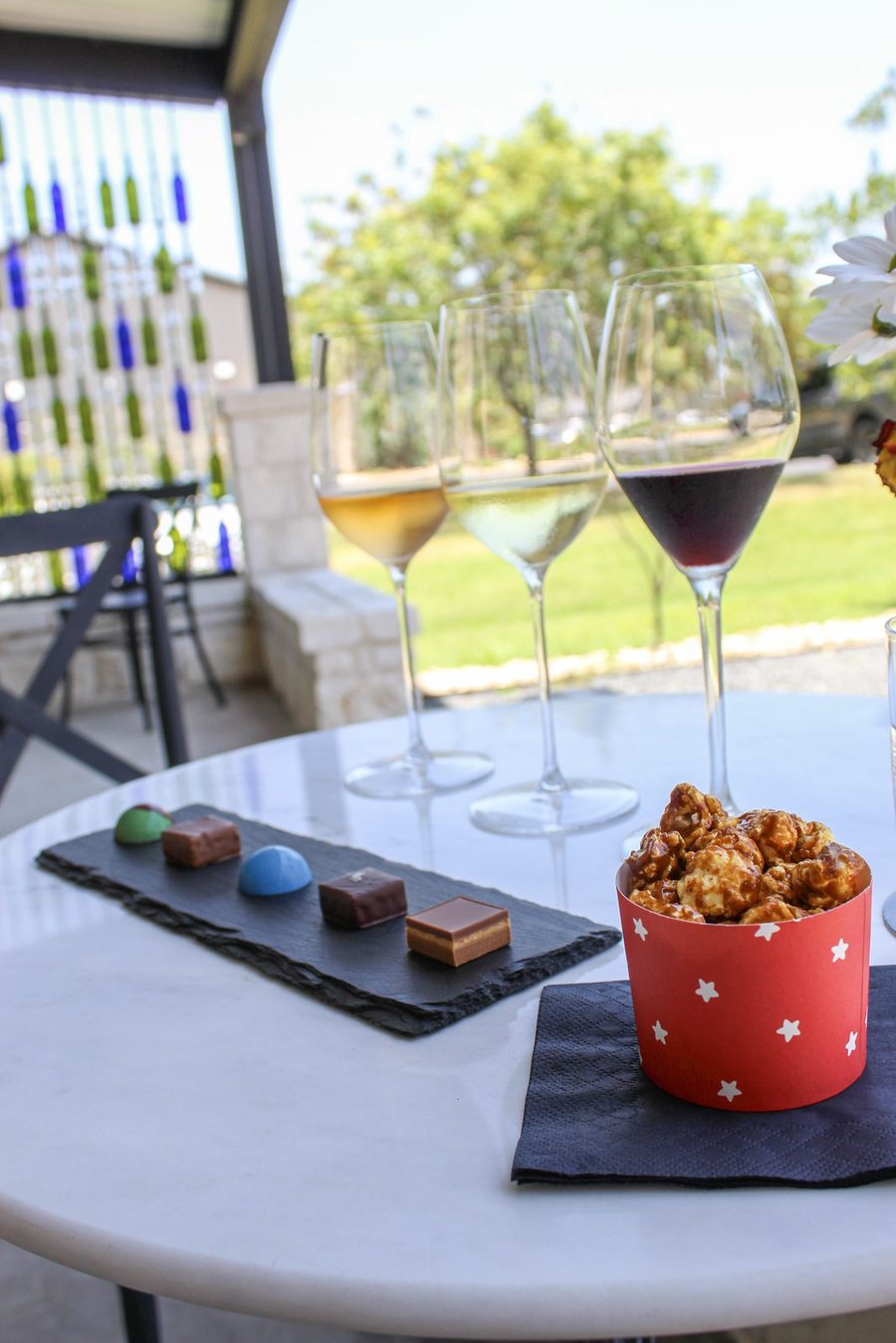 Three glasses of wine on a marble table beside a slate board of five bonbons and a cup of caramel popcorn