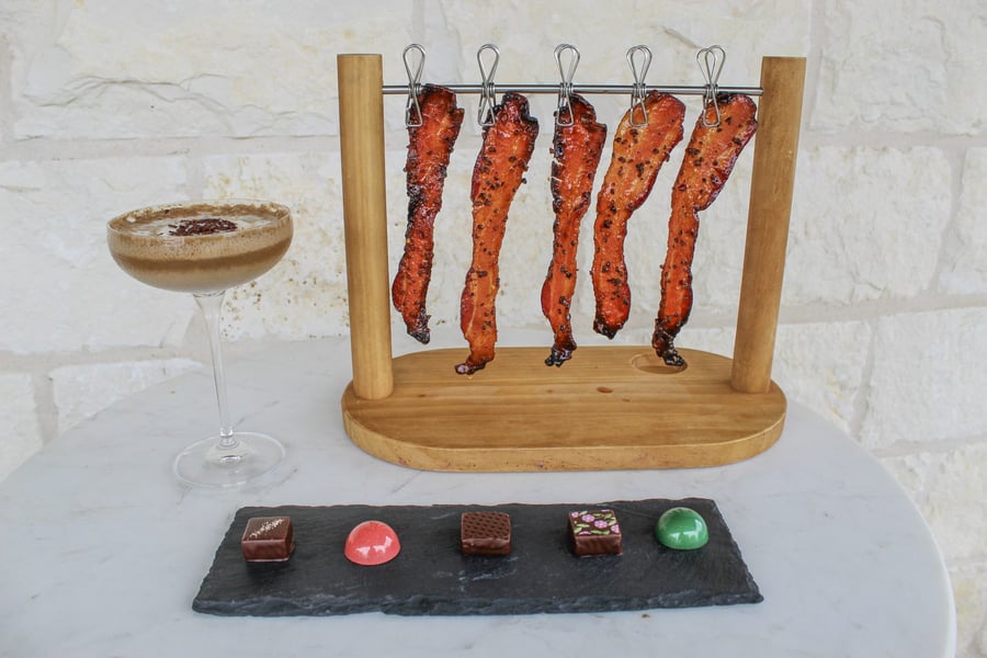 Five glazed bacon strips hanging from a wooden rack beside a chocolate coupe and a slate of bonbons