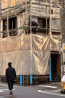 Scaffolding for a building construction with a worker sitting on it working and a passerby looking towards it. Sun casting on the tarp showing it was in the afternoon