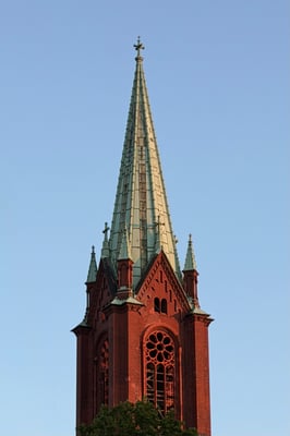 a tall ornamented brick church with a pointed roof in a shade of oxidized copper (greenish cyan).