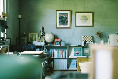 a room with a bass guitar and bookshelves, with paintings hung with wooden frames on a concrete wall, with natural light coming from one side of the room.