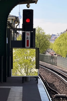 an overground train tracks next to a stop light with a mirror reflecting trees on the street, Berlin tv tower can be seen in the background