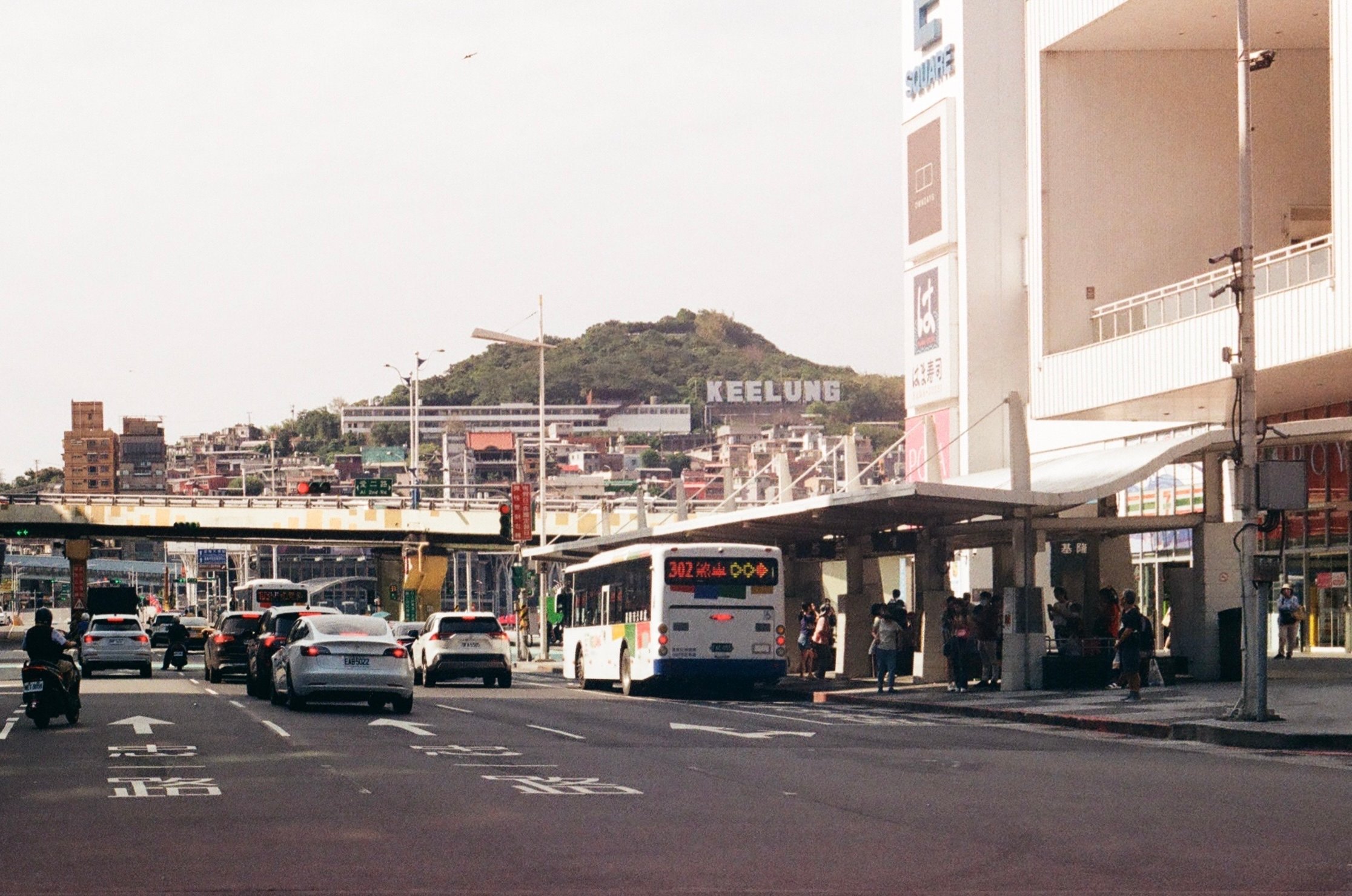 a bus and cars at a bus stop with a sign in the background on a hill reading KEELUNG