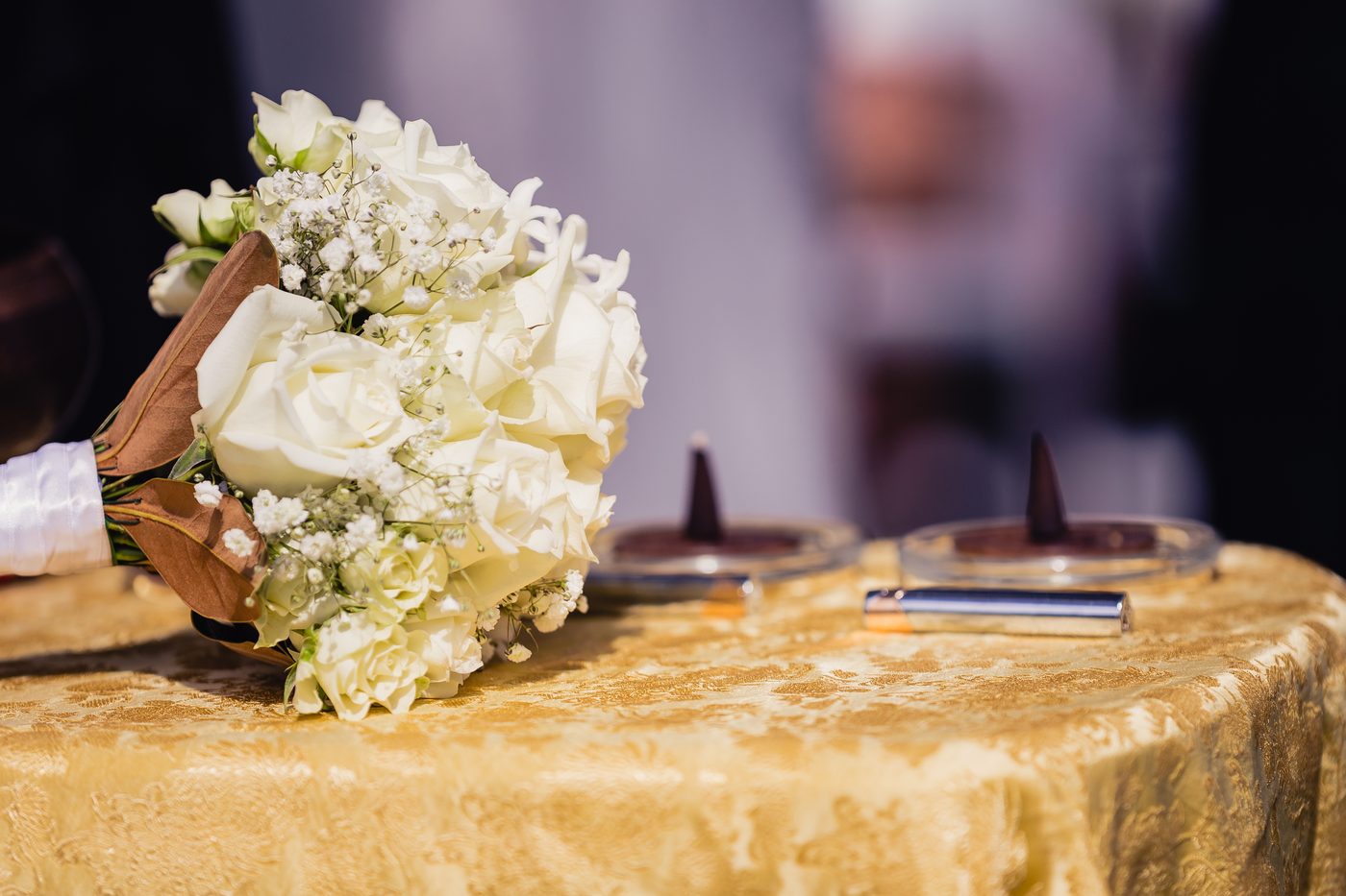A bridal bouquet of white roses and baby's breath rests on a gold-patterned tablecloth at the, with two small candles and a silver lighter also on the table. Engagement ceremony by She Said Yes.