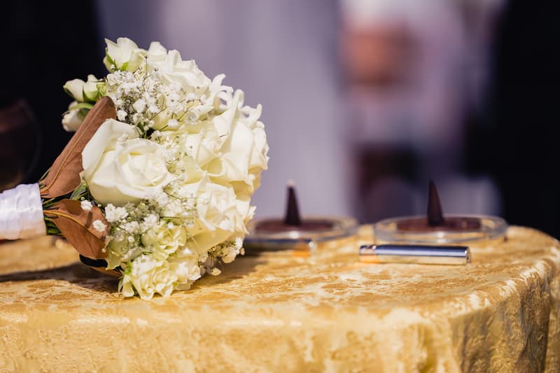 A bridal bouquet of white roses and baby's breath rests on a gold-patterned tablecloth at the Toowong Rowing Club, with two small candles and a silver lighter also on the table.