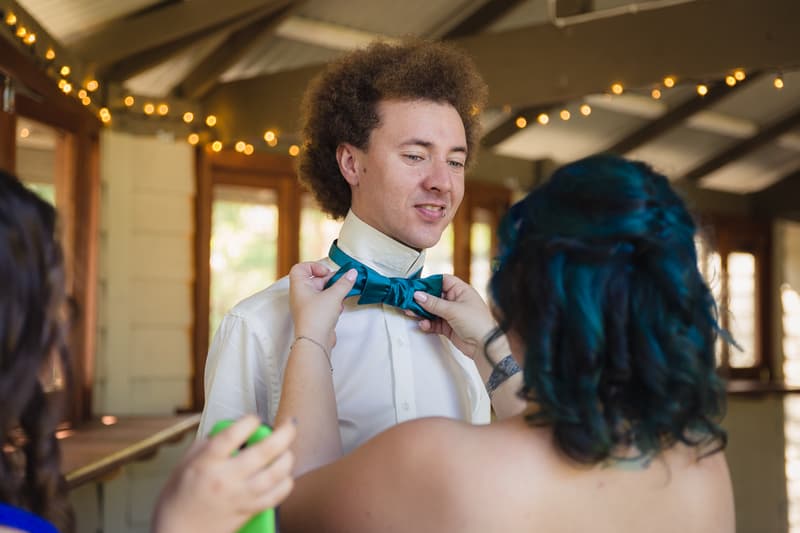 A bridesmaid adjusts the groom Paul's teal bow tie inside Kwila Lodge while he stands wearing a white shirt.