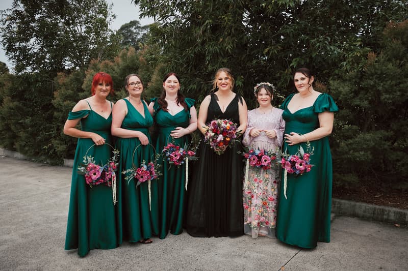 The bride Jayme in a black dress stands with five bridesmaids in green dresses and a flower girl in a floral dress holding bouquets at Sandstone Point Hotel — The Pavilion.