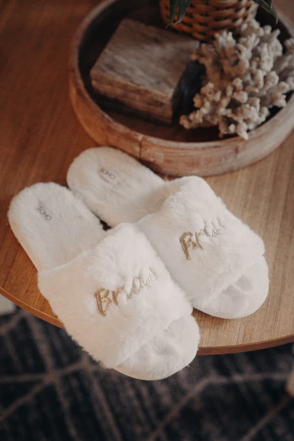 A pair of white fluffy slippers embroidered with the word 'Bride' placed on a wooden table with a decorative wooden bowl containing dried flowers.