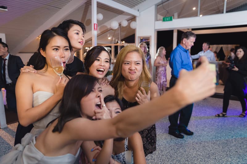 A group of six female wedding guests pose closely together for a selfie at the reception stage of Bilinga Beach Weddings — Bilinga SLSC, with some holding drinks and making playful faces.