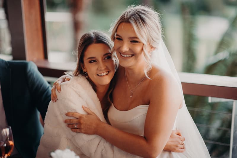 The bride Libby in a strapless wedding gown and veil hugs a bridesmaid wrapped in a white shawl at The Tides — The Pandanus Room during the reception.
