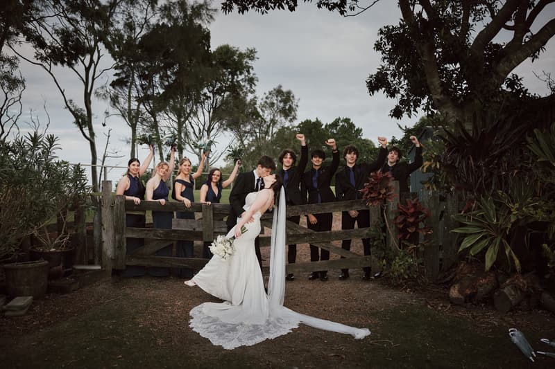 Terion and Tyler share a kiss in front of a wooden gate with bridesmaids and groomsmen standing behind it, all raising their arms in celebration at Sandstone Point Hotelallo.
