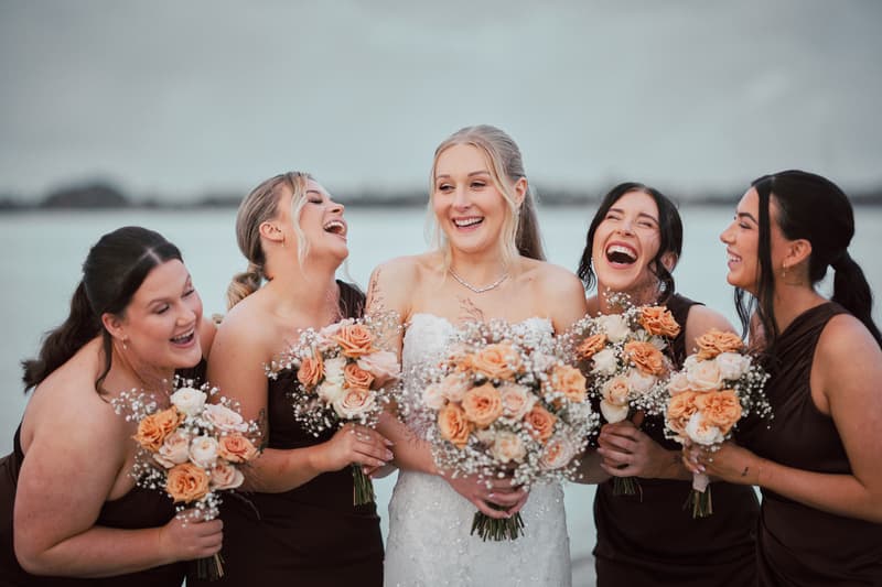 The bride Morgan stands with four bridesmaids holding bouquets of peach and white roses with baby's breath, all laughing together outdoors near water at Sandstone Point Hotel.