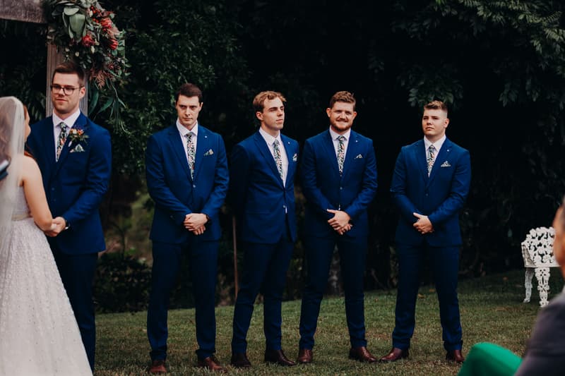 Connor stands with his groomsmen in blue suits with floral ties at Yabbaloumba Retreat — By The River during the wedding ceremony, facing Lilly who is partially visible in her wedding dress and veil.