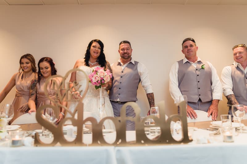 Bride Kelly and groom Shane stand together behind a table at Novotel Sunshine Coast Resort reception, flanked by bridesmaids and groomsmen dressed in matching attire, with a decorative 'So hitched' sign in the foreground.