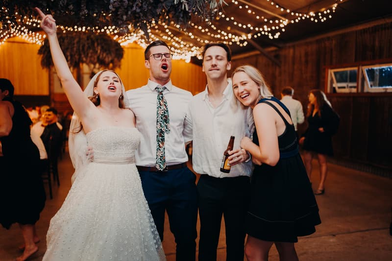 The bride Lilly, the groom Connor, and two guests sing and celebrate together at the reception stage in The Shed at Yabbaloumba Retreat, with string lights overhead.