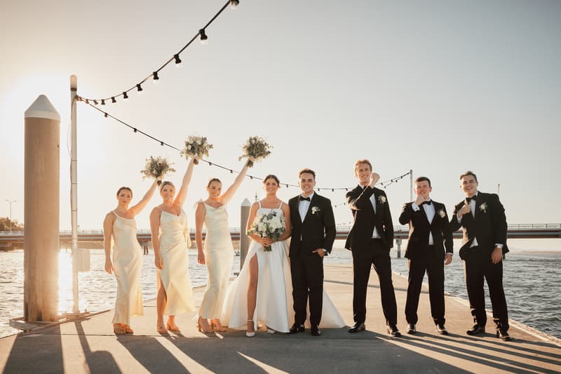 Ashleigh in her wedding gown and James in a tuxedo stand together on a pier at Sandstone Point Hotel with their bridal party, including four bridesmaids in matching light-colored dresses holding bouquets and three groomsmen in tuxedos.