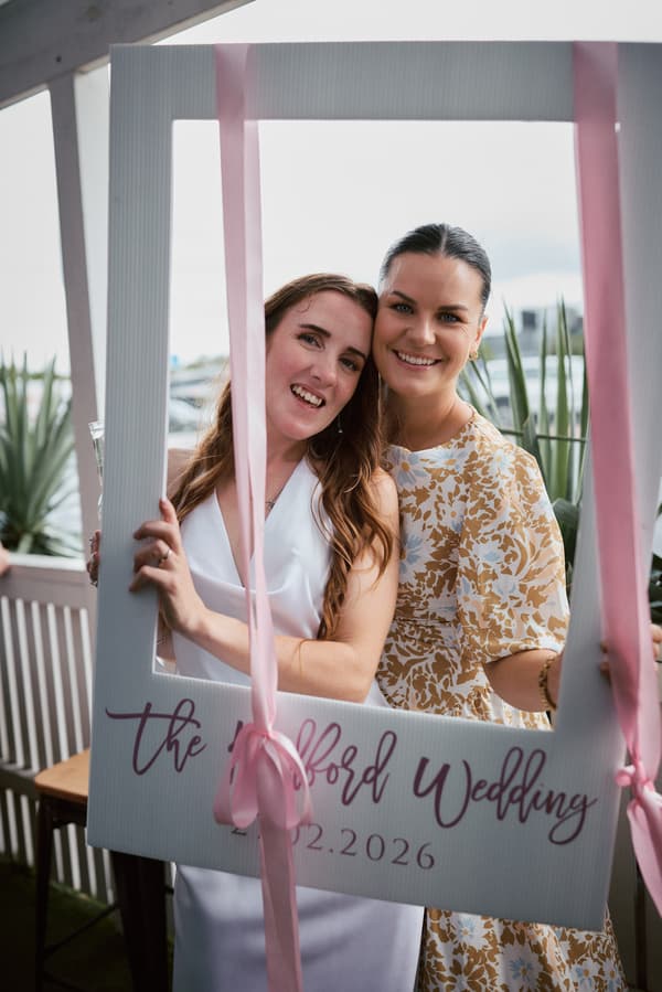 The bride Olivia and a female guest pose together holding a large photo frame prop at the White Horse Ranch reception.