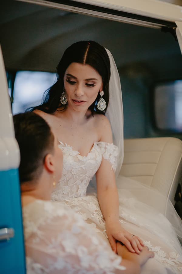 The bride in a white lace off-shoulder wedding dress and veil sits inside a vehicle, holding hands with another person wearing a lace outfit.