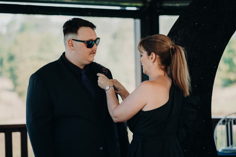 A woman adjusts the boutonniere on a man wearing sunglasses and a dark suit at Ocean View Estates — On The Lake.