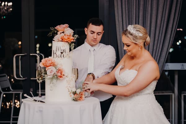 Emily the bride and Dylan the groom cut their wedding cake decorated with pink and peach flowers at Sandstone Point Hotel — Pumicestone Room.
