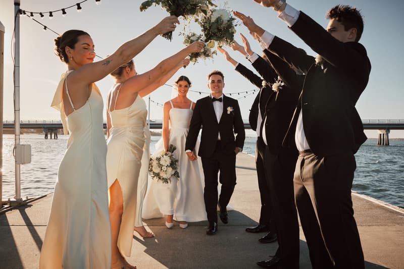 Bride Ashleigh and groom James walk hand in hand under an arch formed by bridesmaids and groomsmen holding bouquets and raising their arms at Sandstone Point Hotel pier.